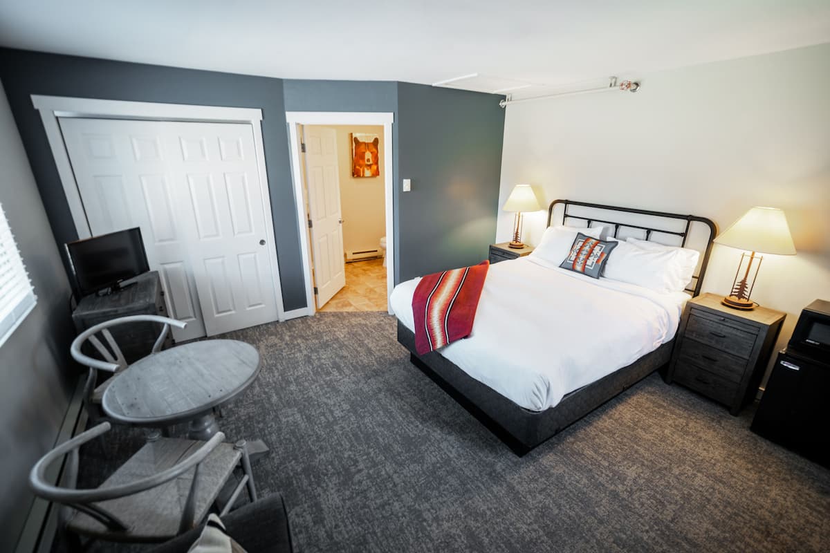 Une chambre d’hôtel comprenant un lit, deux tables de chevet avec des lampes, une petite table ronde avec des chaises, un téléviseur et une porte donnant sur une autre pièce.