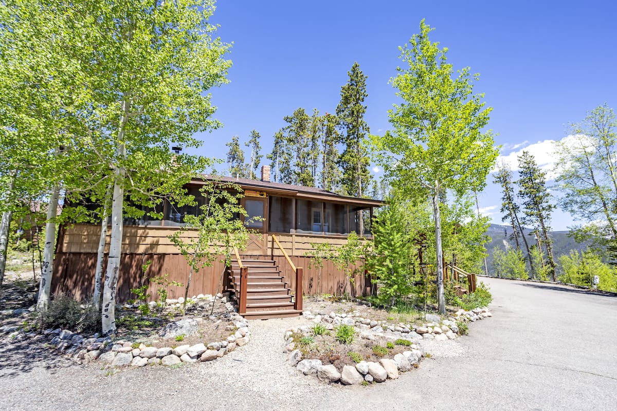 Une cabane en bois à deux étages, avec un escalier menant à une terrasse, entourée d’arbres et bordée d’un chemin de gravier.