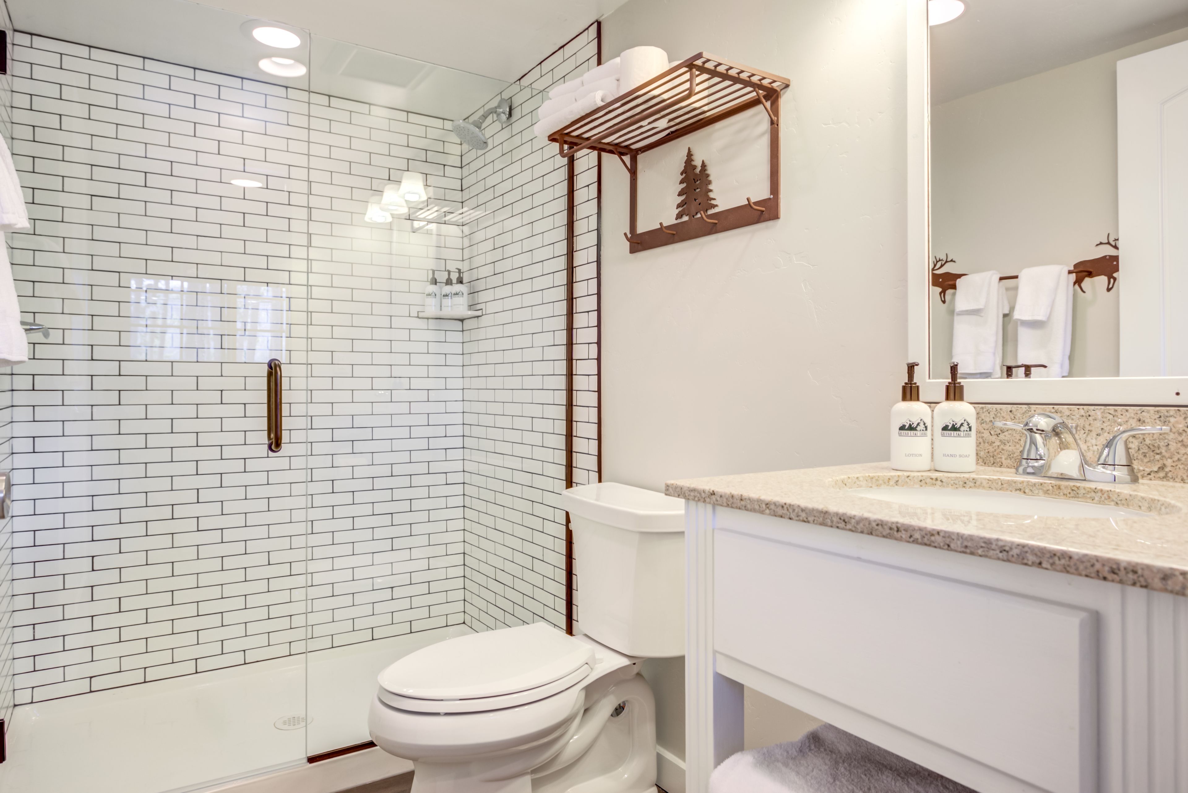 A modern bathroom with a glass shower enclosure, white tiled walls, a toilet, a sink with a granite countertop, and a mirror with a towel rack.