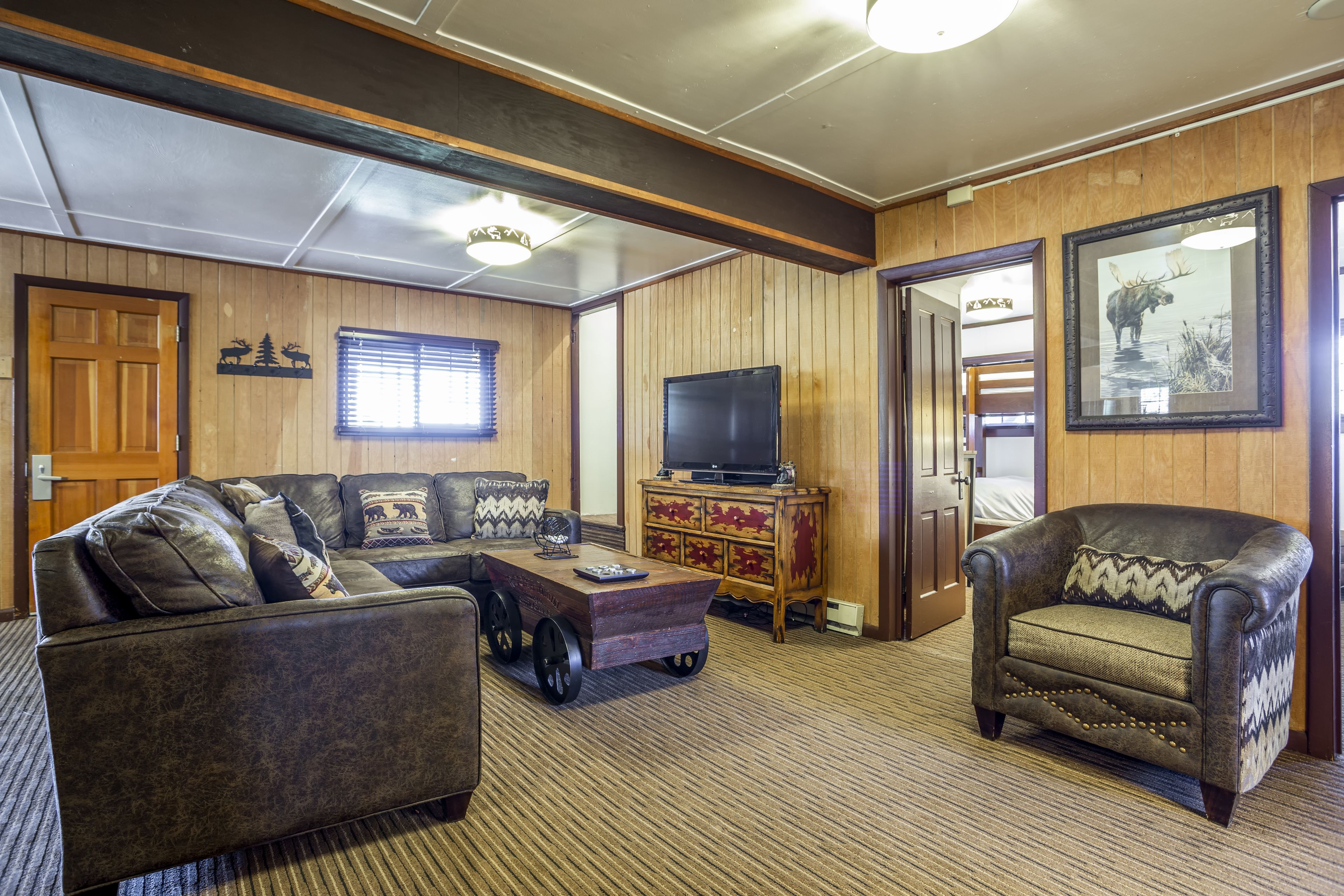 A cozy living room with a sectional sofa, a small armchair, a wooden cabinet with a television, and a door leading to another room.