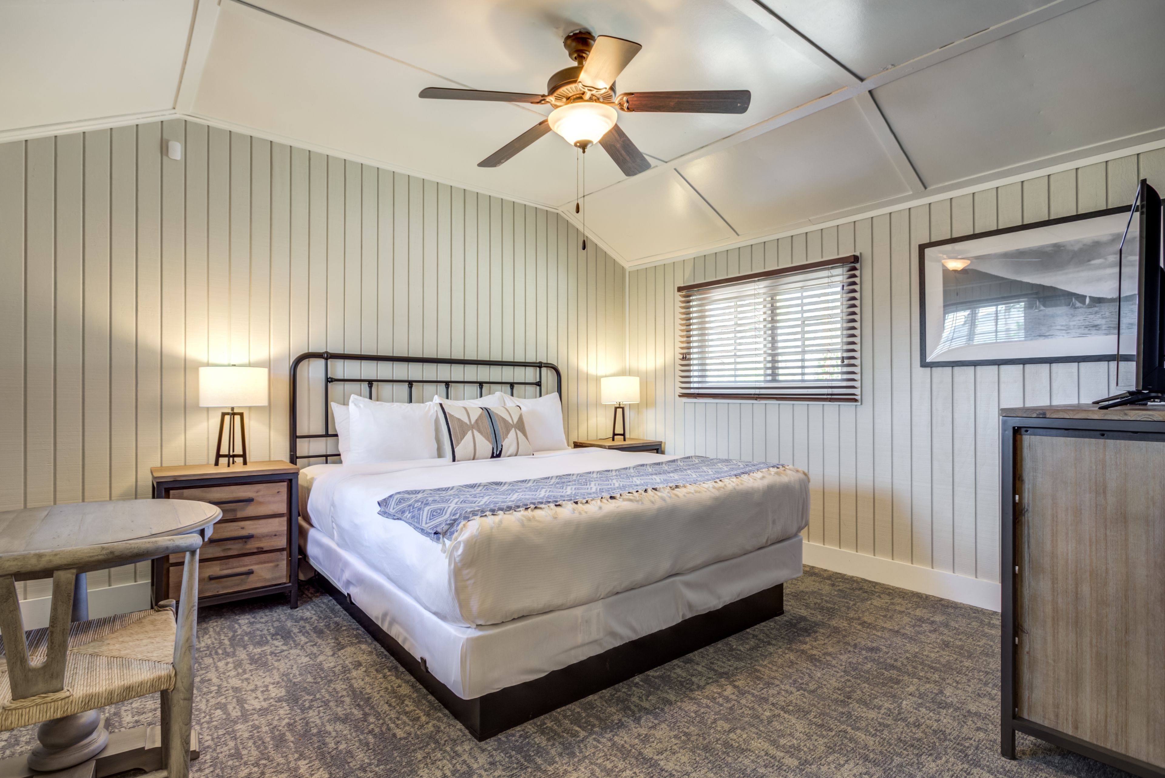 A bedroom with a bed, bedside tables, a ceiling fan, a window with blinds, and a framed picture on the wall.