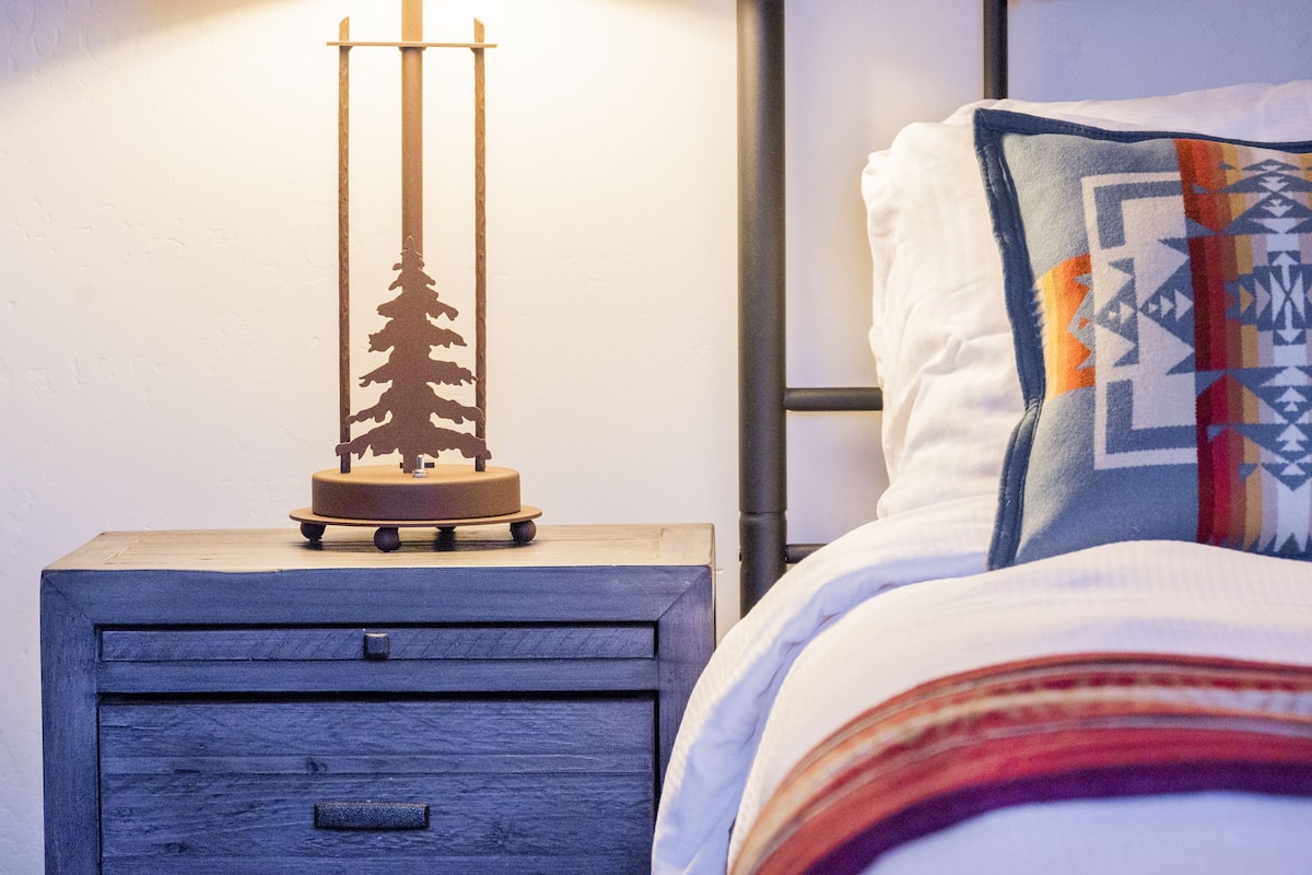 Une table de chevet en bois avec un tiroir, un objet décoratif et un lit agrémenté d’un coussin à motifs.