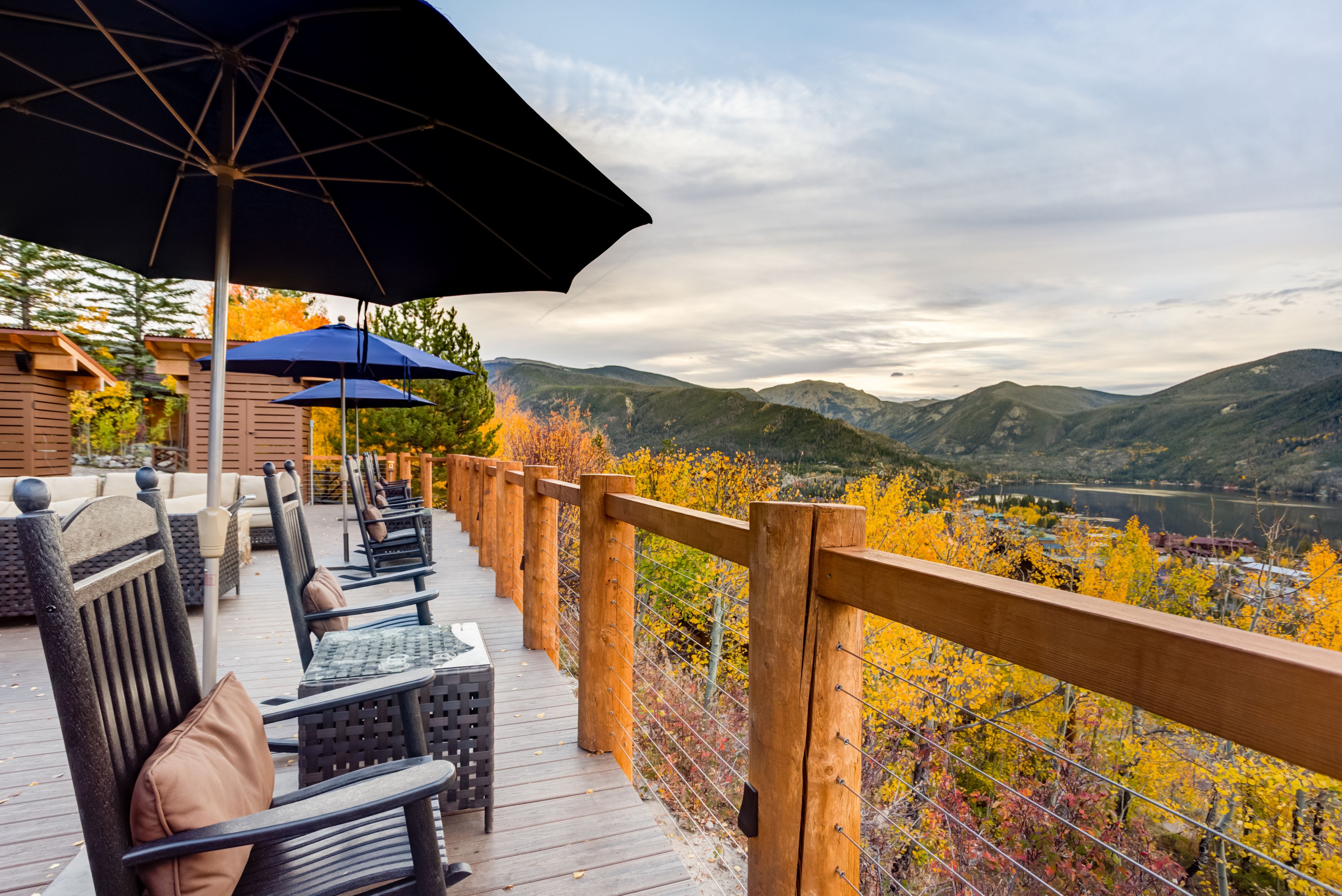 A wooden deck with outdoor seating and umbrellas, overlooking a mountainous landscape with autumn foliage.