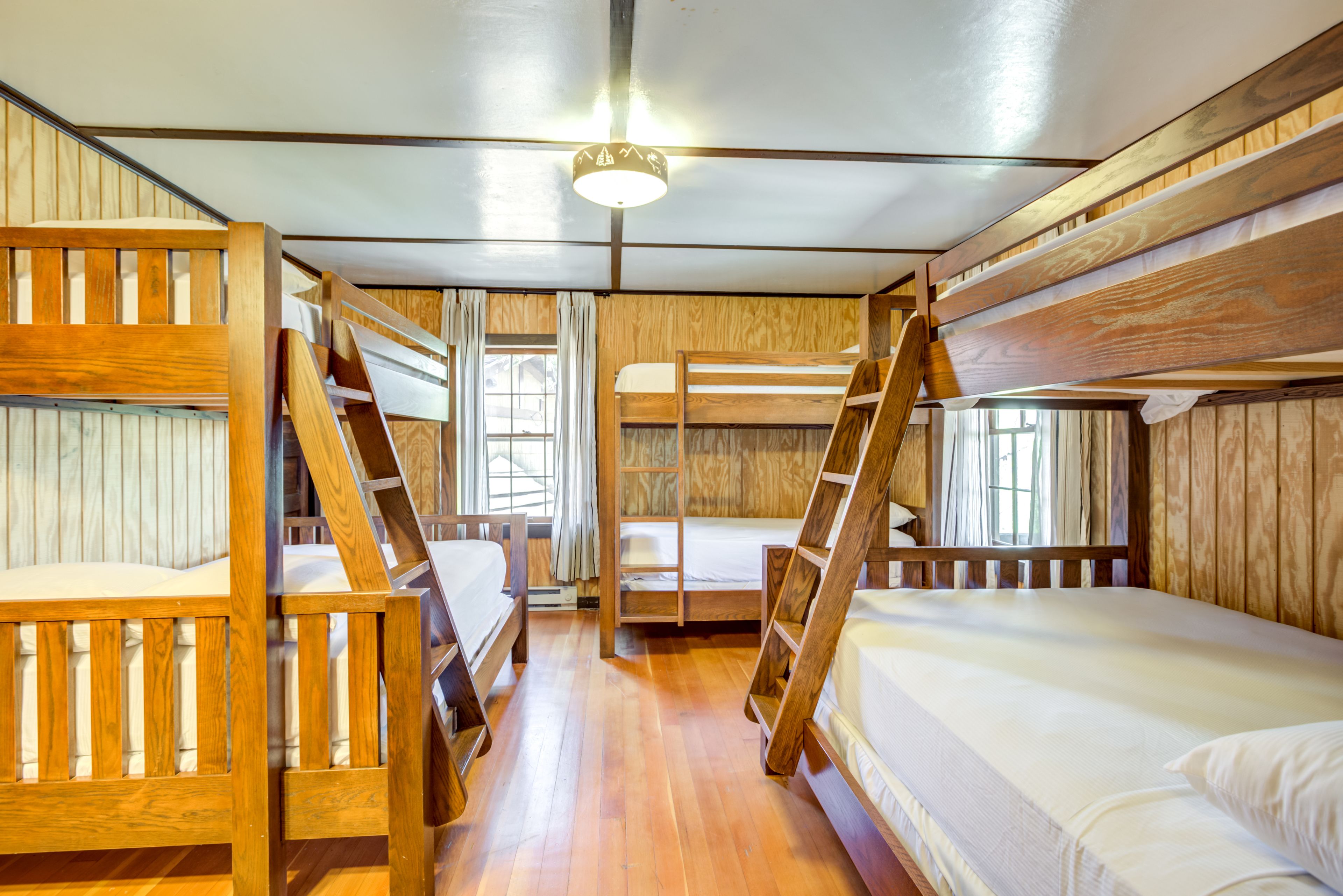 A bunk bed room with wooden walls and floors, a ceiling light, and a window with curtains.