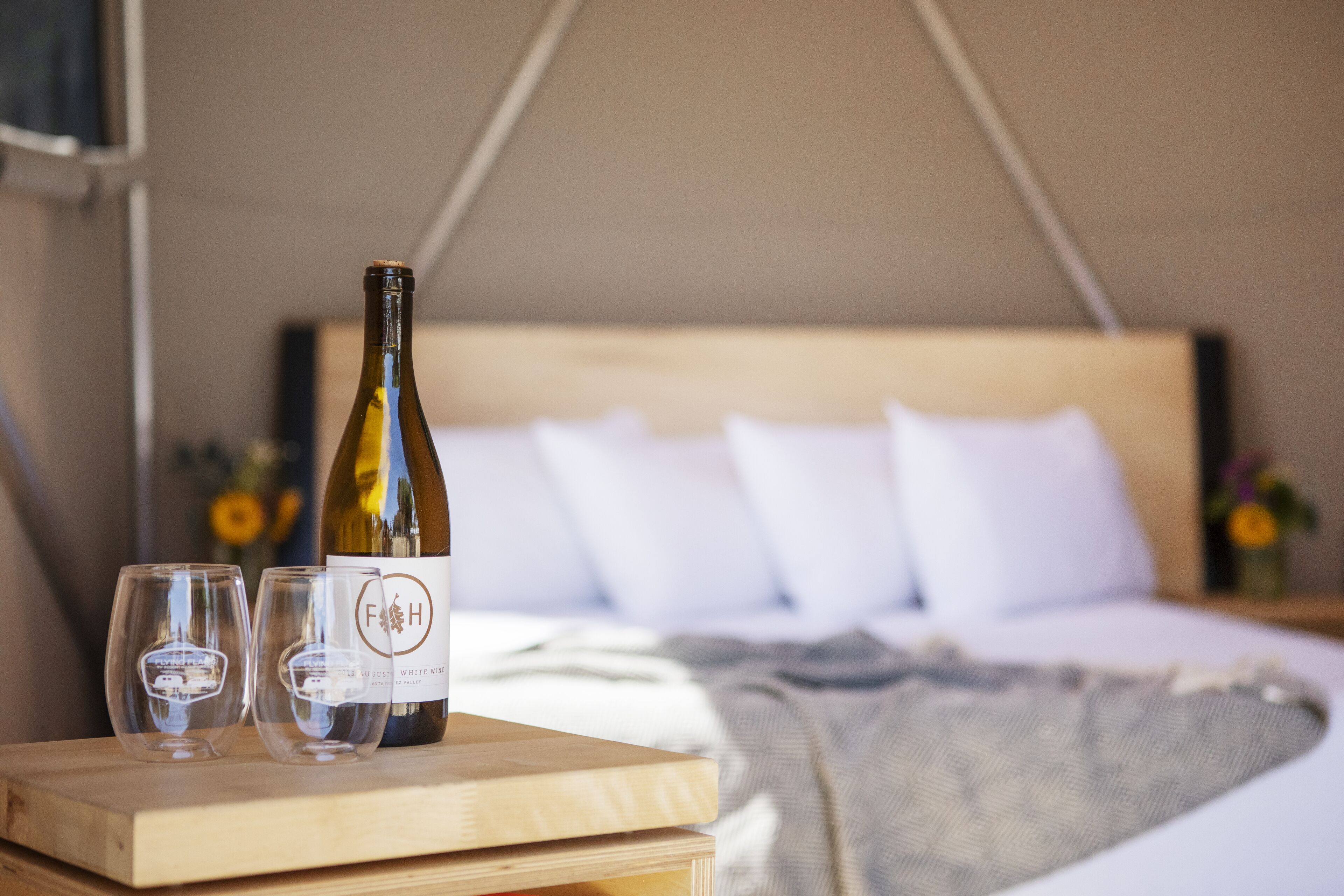 A bed with white pillows and a wooden headboard, a bedside table with a wine bottle and two glasses, and a blanket on the bed.
