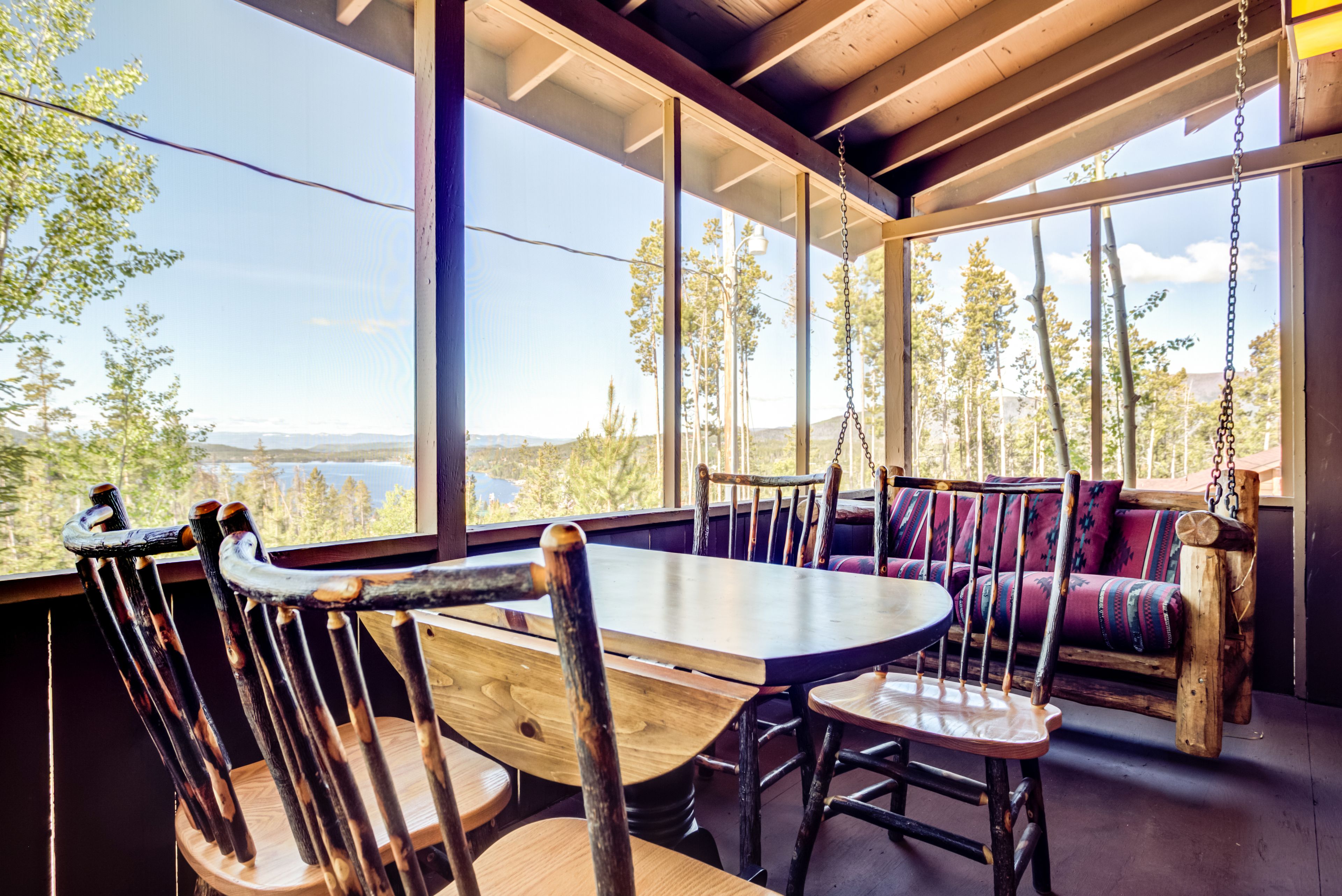 Una mesa de comedor de madera con sillas, un sofá y un columpio colgante en una habitación con ventanas amplias que dan a un bosque y un lago.