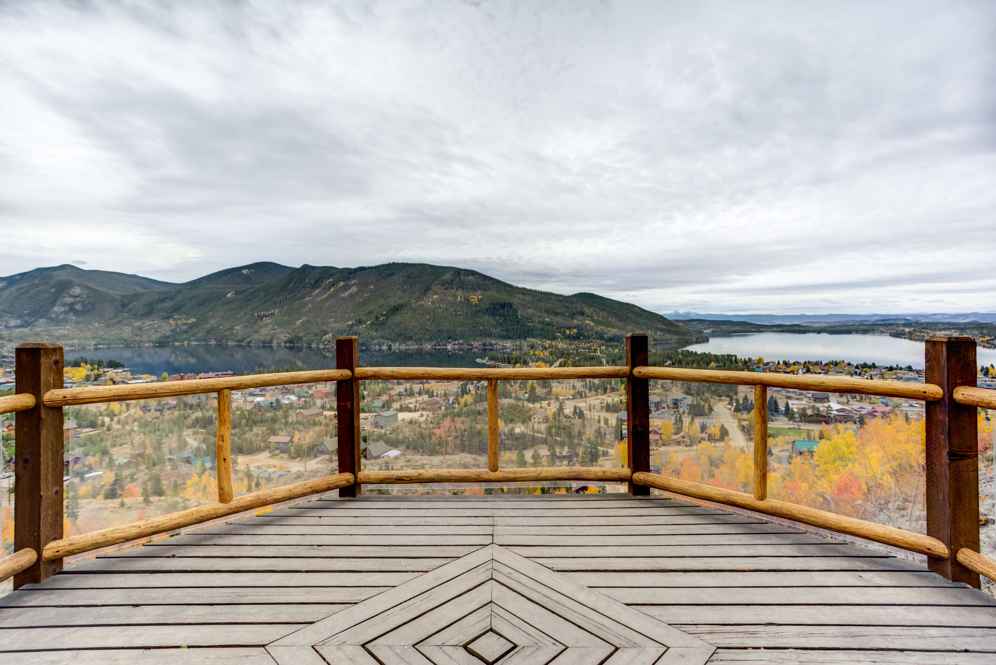 Una terraza de madera con un diseño geométrico, con vistas a un paisaje montañoso con un lago y árboles de colores otoñales.