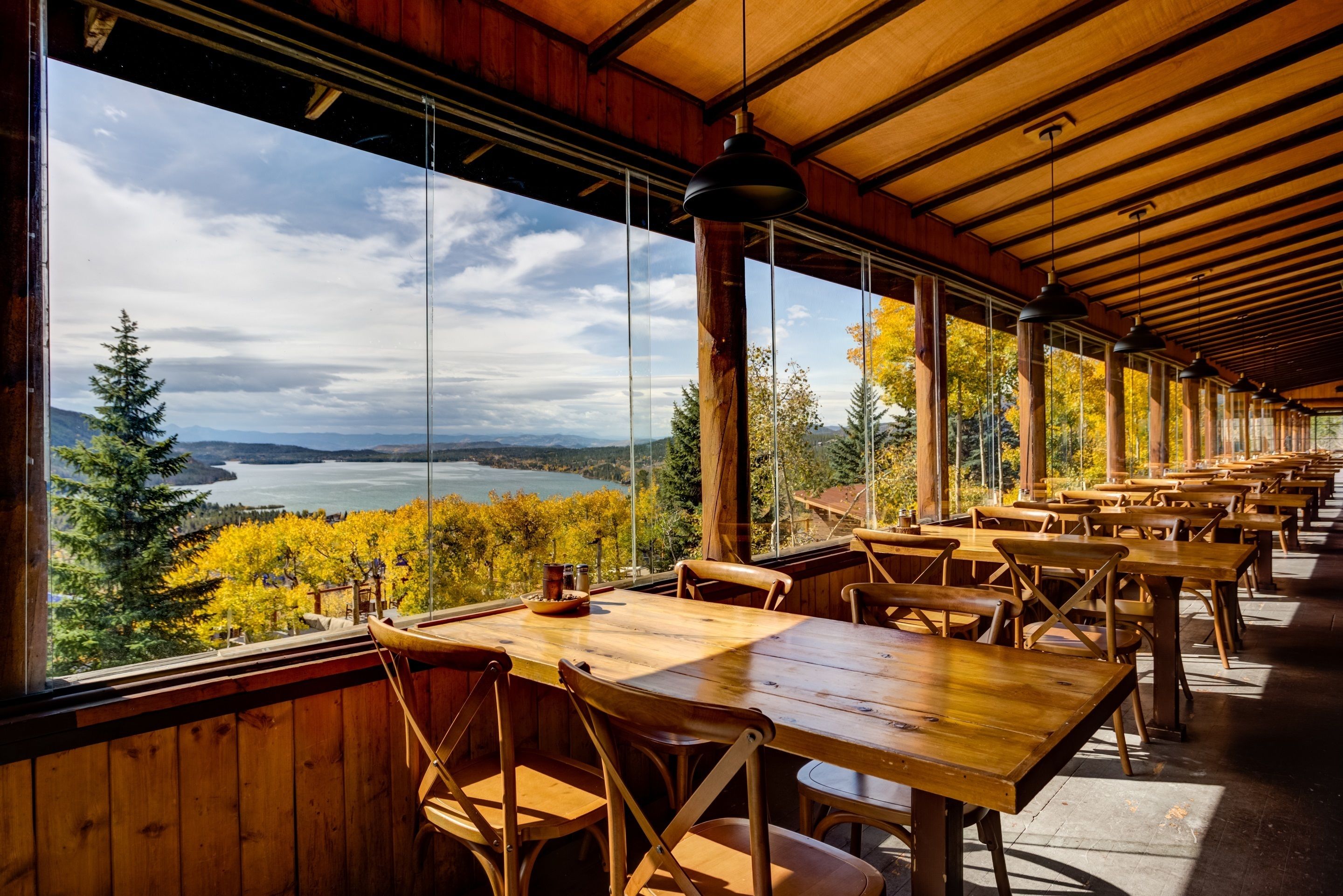 Een restaurant met houten tafels en stoelen, grote ramen met uitzicht op een meer en herfstkleurige bomen.