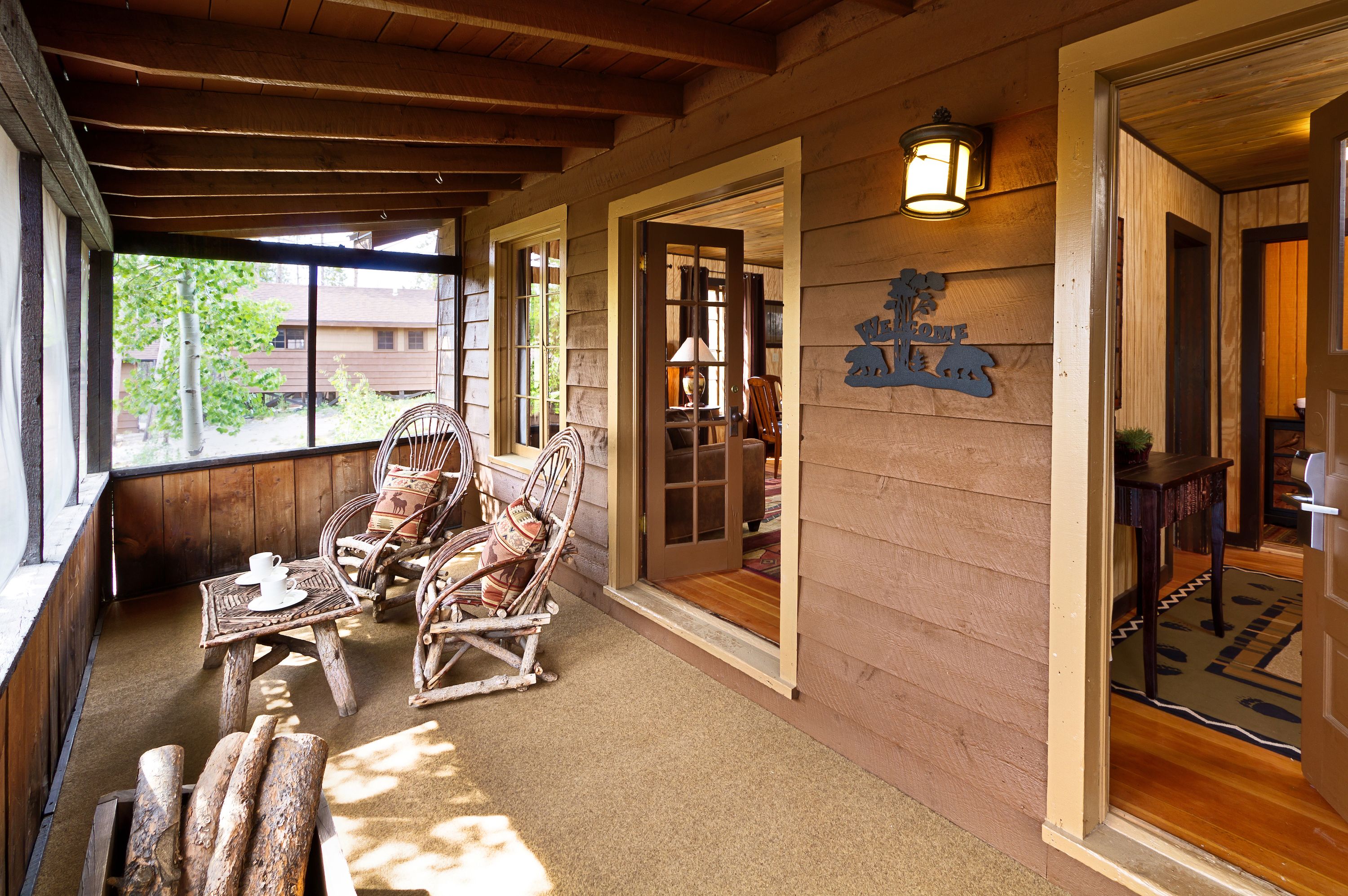 A wooden porch with wicker chairs and a table, a wooden door with a sign, and a view of a building through a window.