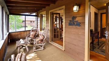 A wooden porch with wicker chairs and a table, a wooden door with a sign, and a view of a building through a window.