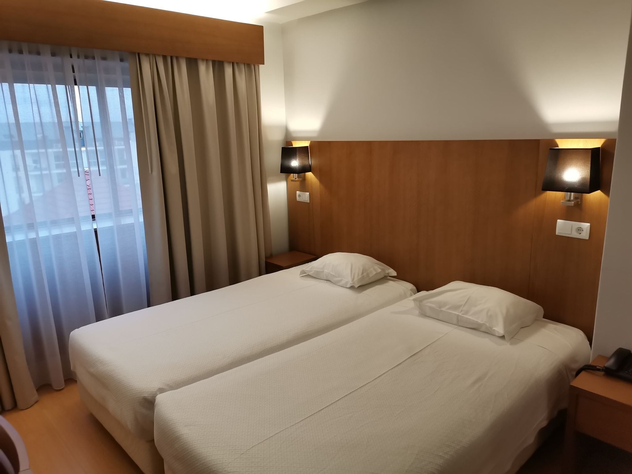 Une chambre d’hôtel avec deux lits, une tête de lit en bois, une table de chevet avec une lampe et une fenêtre avec des rideaux.