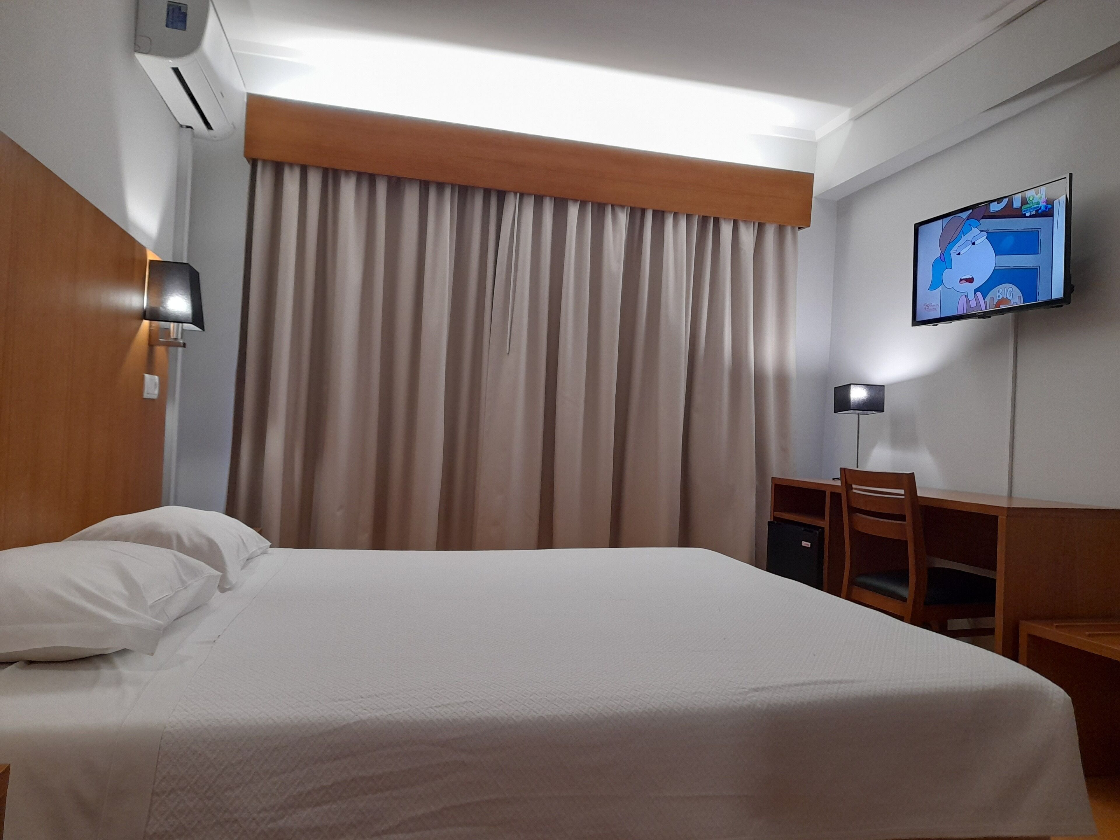 A hotel room with a bed, a TV mounted on the wall, a desk with a chair, and a window with curtains.