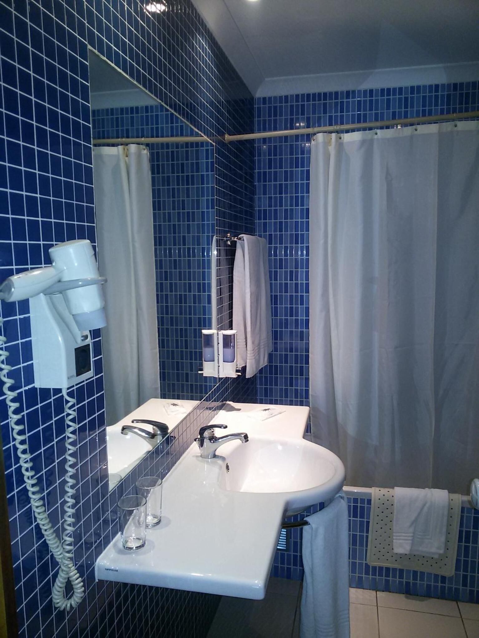Een badkamer met blauwe betegelde wanden, een witte wastafel, een spiegel, een haardroger en een douchegordijn.