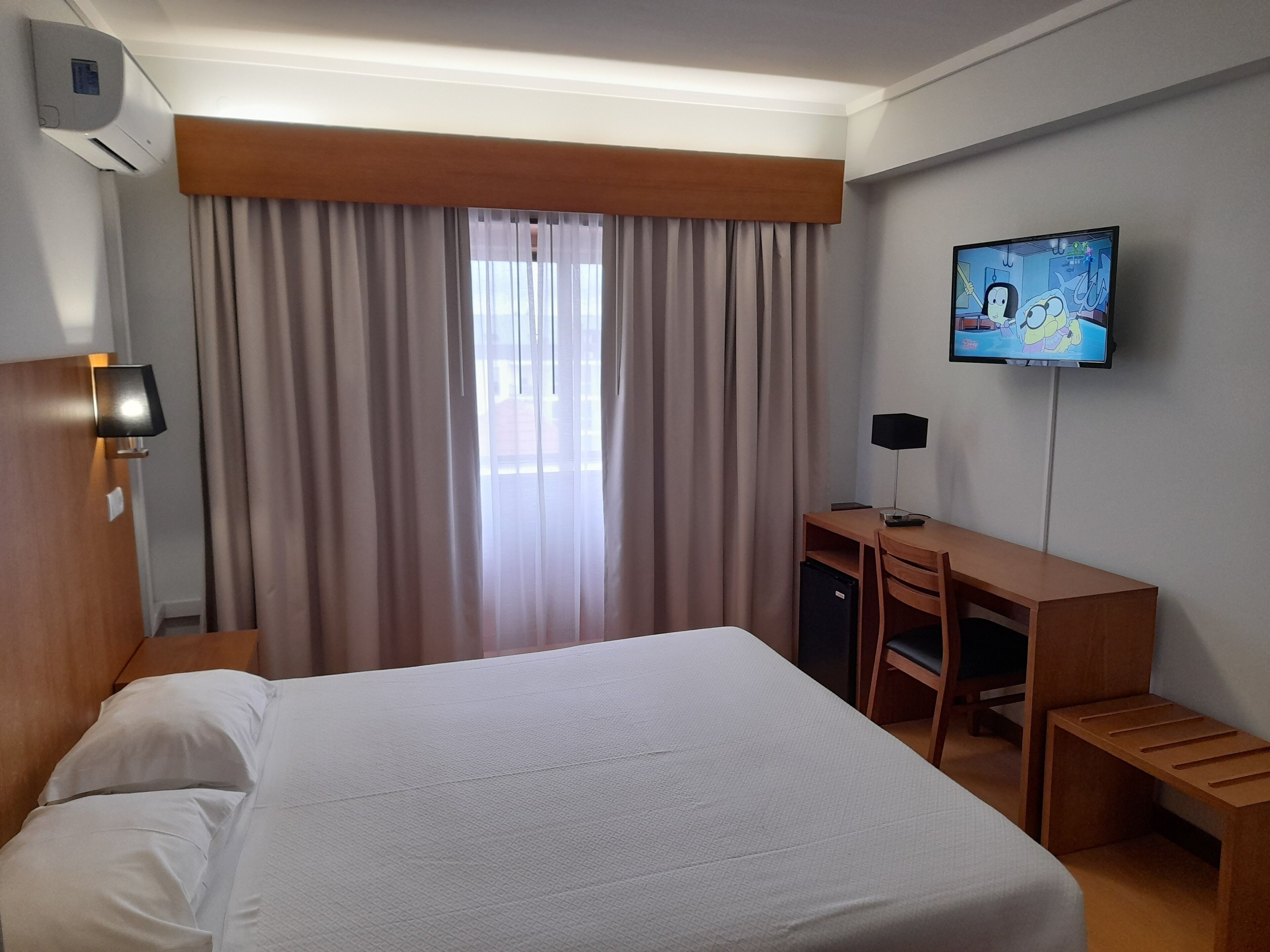 Habitación de hotel con cama, escritorio, televisor y ventana con cortinas.