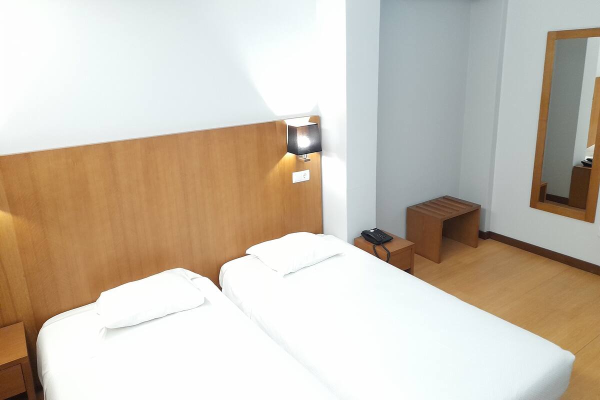A hotel room with two single beds, a nightstand, a bedside table with a lamp, a small wooden stool, and a mirror on the wall.
