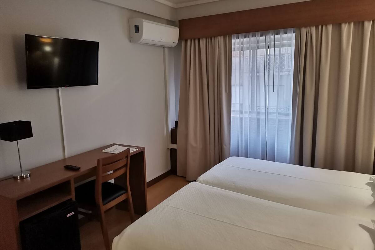A hotel room with a bed, a desk with a chair, a TV, and an air conditioning unit.