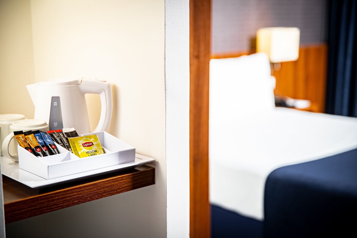 A hotel room with a bedside table holding a coffee maker, a tray with coffee packets, and a small box.