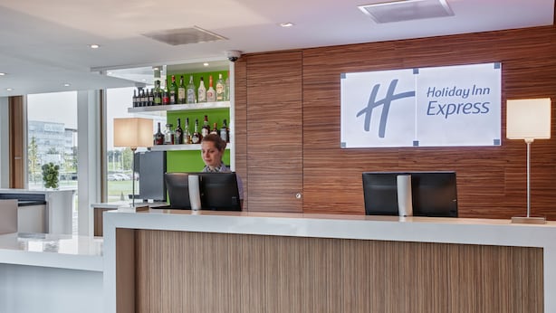 A Holiday Inn Express reception desk with a wooden panel design, a bar area with liquor bottles, and a large sign with the hotel name. - Holiday Inn Express Utrecht - Papendorp by IHG (Utrecht)