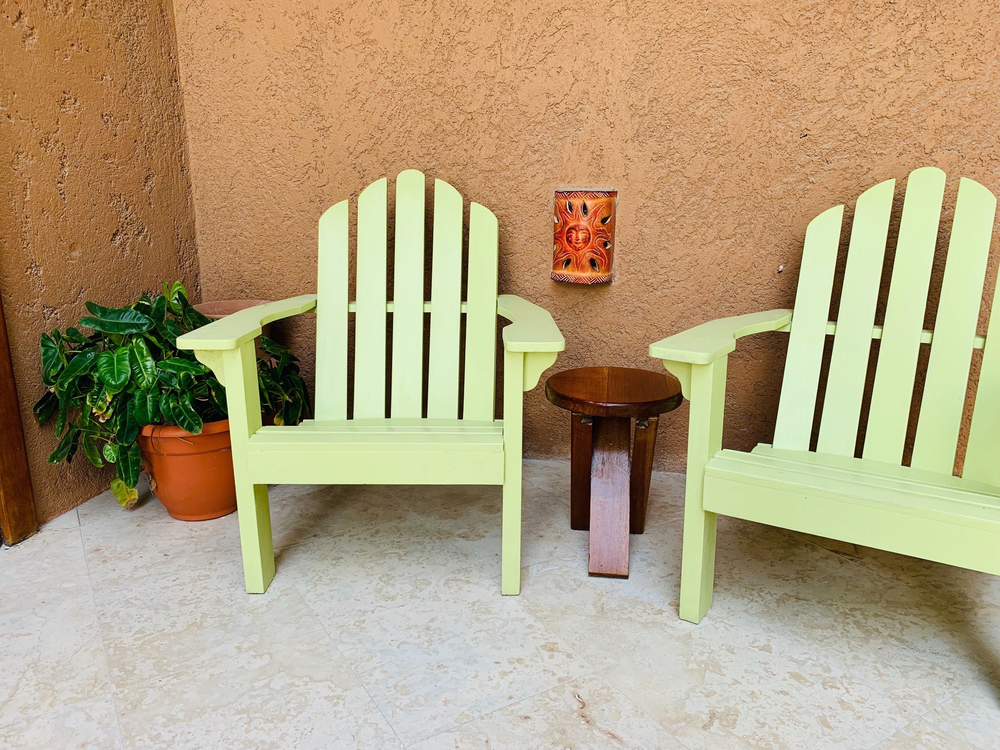 Two light green Adirondack chairs with a small round wooden table between them, placed on a tiled floor against a textured wall.