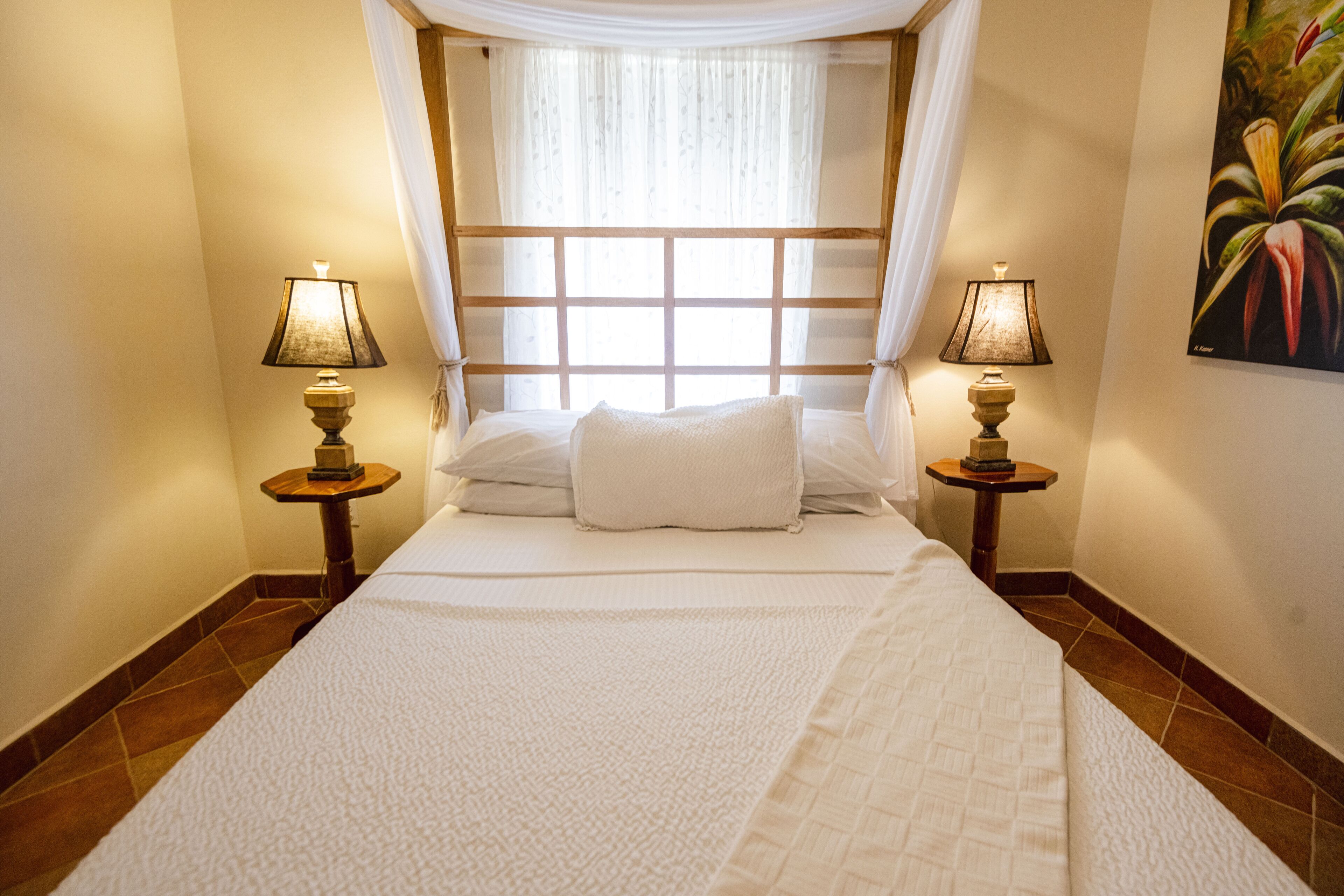 Un letto rifatto con un piumone bianco e cuscini, affiancato da due comodini con lampade, una finestra con tenda e un quadro appeso al muro.