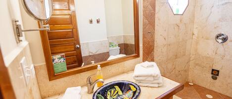 Un baño con tocador de madera, lavabo con un recipiente decorativo, espejo y zona de ducha.
