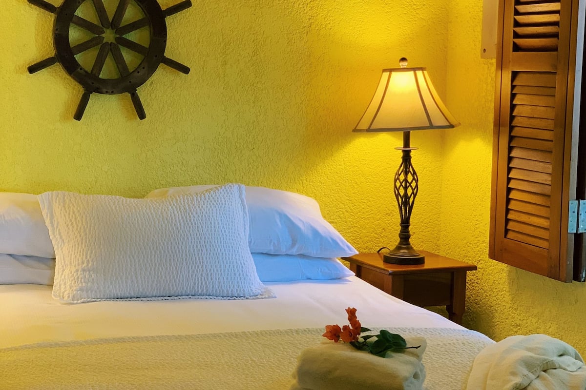 A neatly made bed with white linens, a ship's wheel on the wall, a lamp on a bedside table, and wooden shutters on the window.
