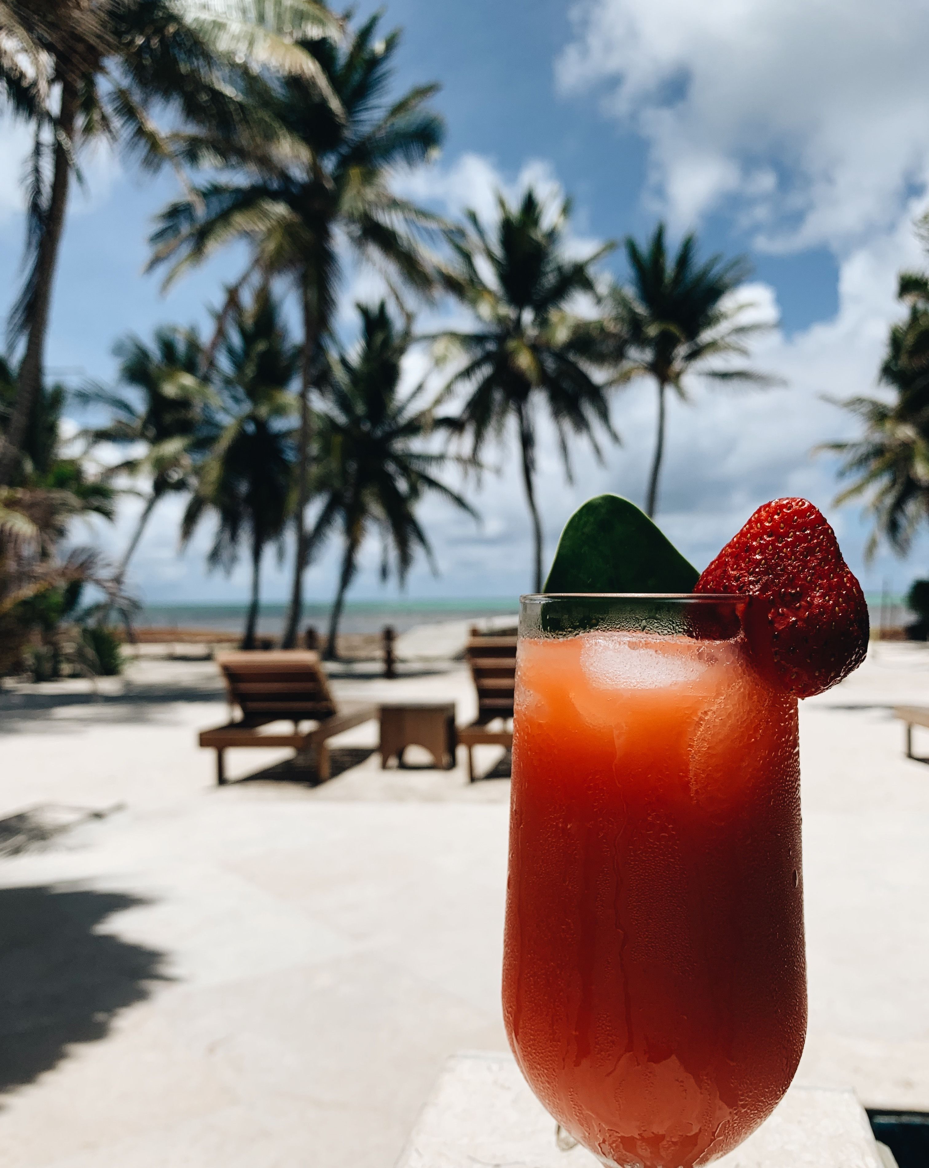 A cocktail glass with a strawberry garnish on a beach with palm trees and lounge chairs.