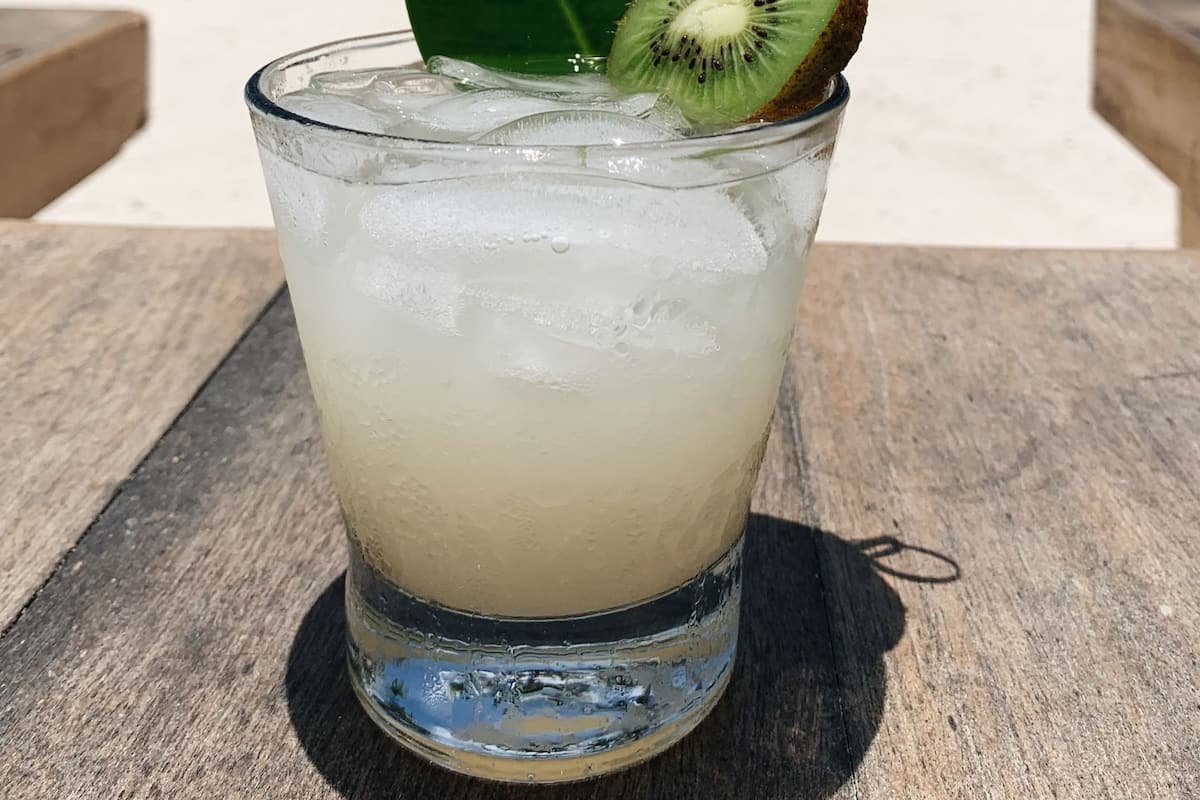 A glass of a light-colored beverage with a slice of kiwi and a leaf garnish on a wooden table at a beach with palm trees and a cloudy sky in the background.