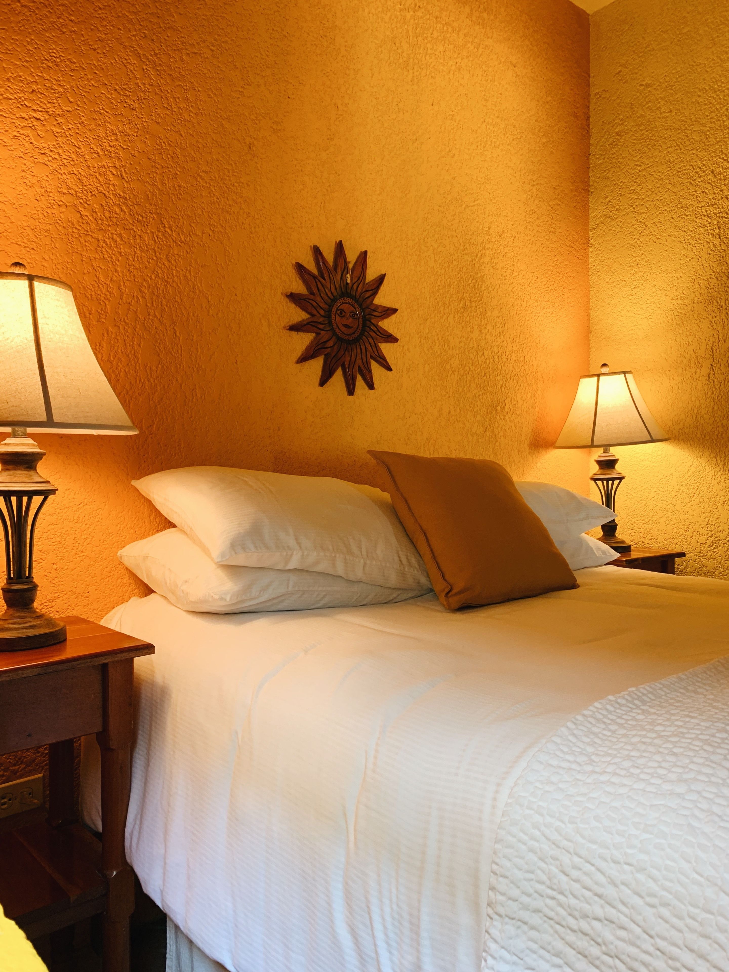 A hotel room with a bed, two bedside lamps, a wall-mounted sun decoration, and a textured wall.
