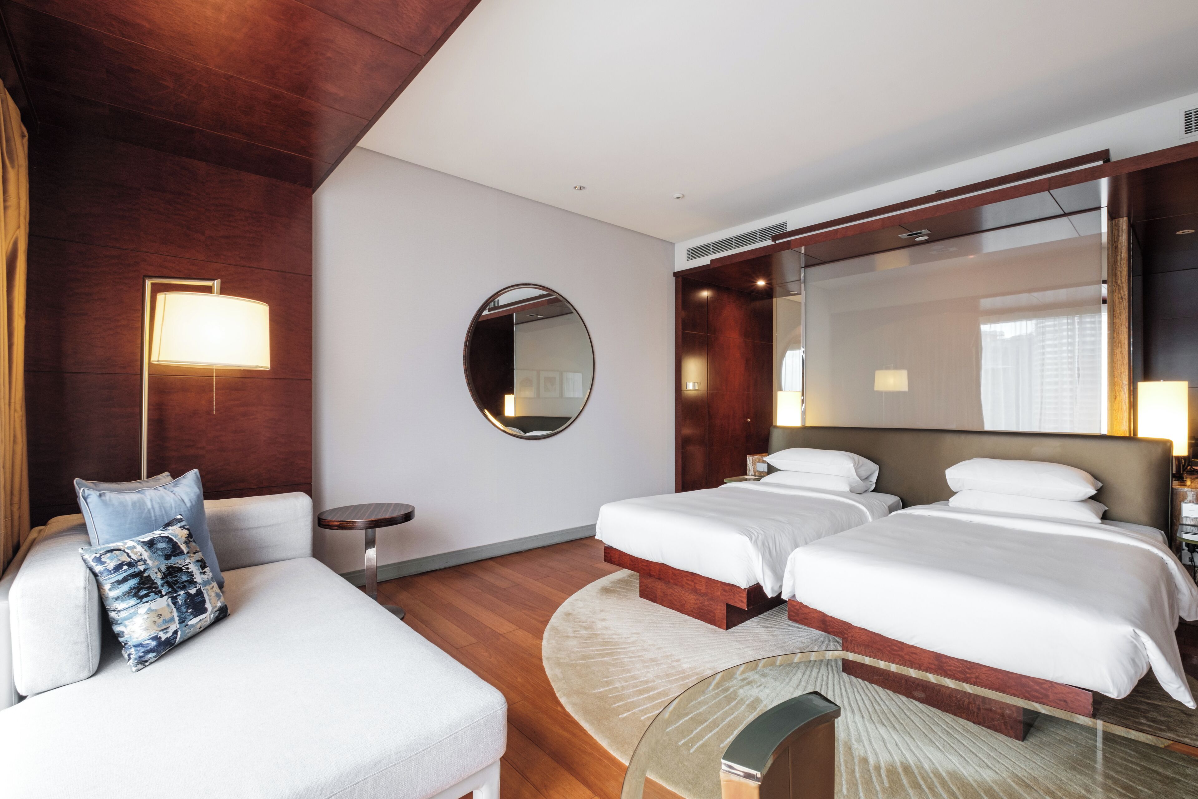 A hotel room with two beds, a sofa, a round mirror, and a small table.