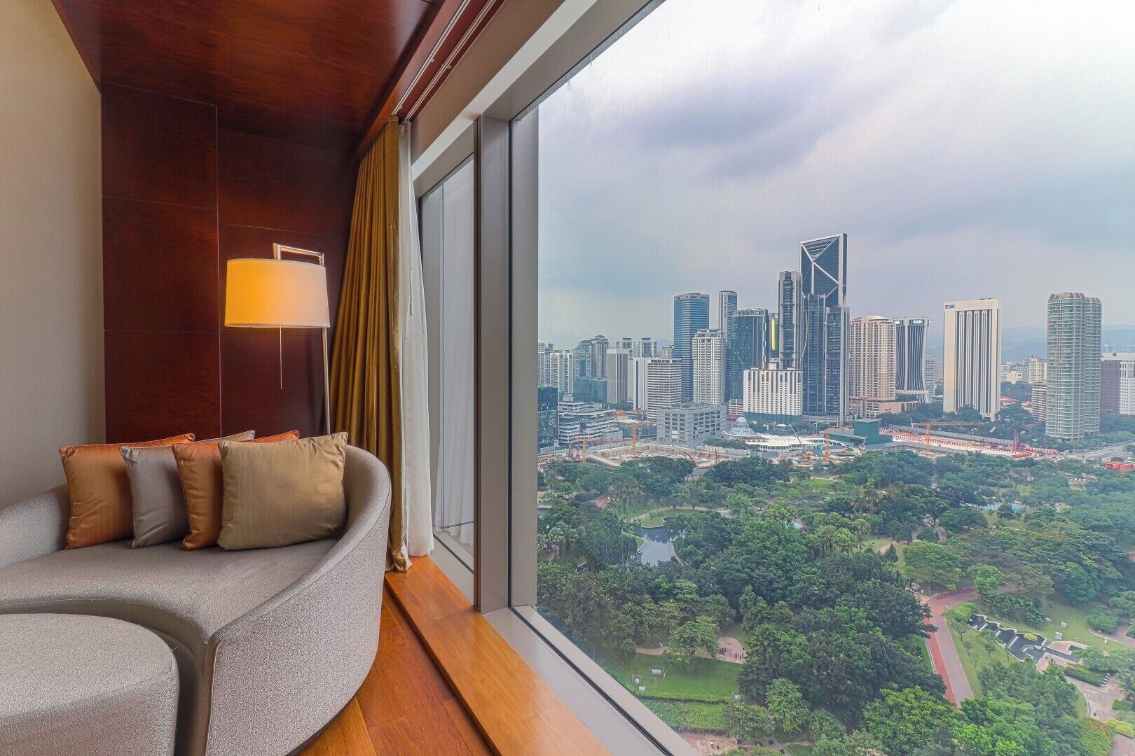 A modern hotel room with a curved sofa, a lamp, and a large window offering a cityscape view.
