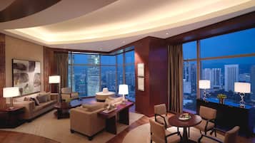 A modern hotel room with a large city view, a sofa, a dining table with chairs, and a desk with a lamp.