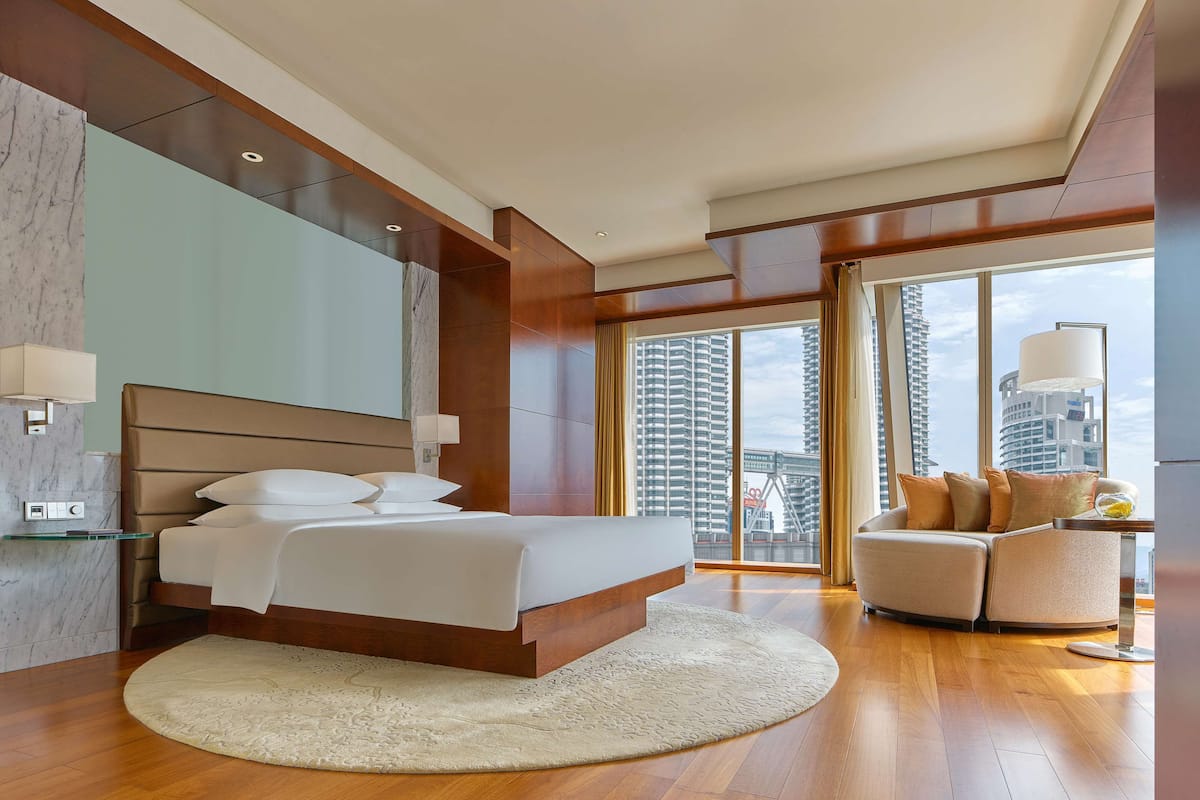 Een moderne hotelkamer met een groot bed, een slaapbank, een rond tapijt en uitzicht op de stadszicht door grote ramen.