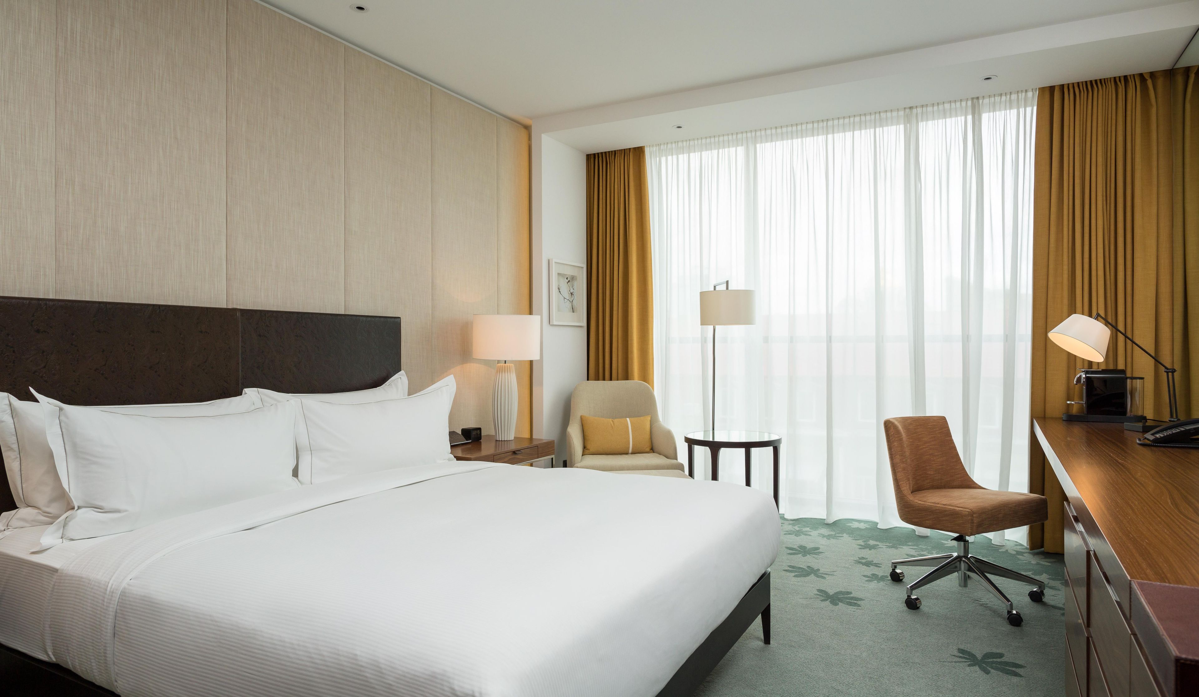 Habitación de hotel con una cama grande, un escritorio con silla, una lámpara y un ventanal con cortinas.