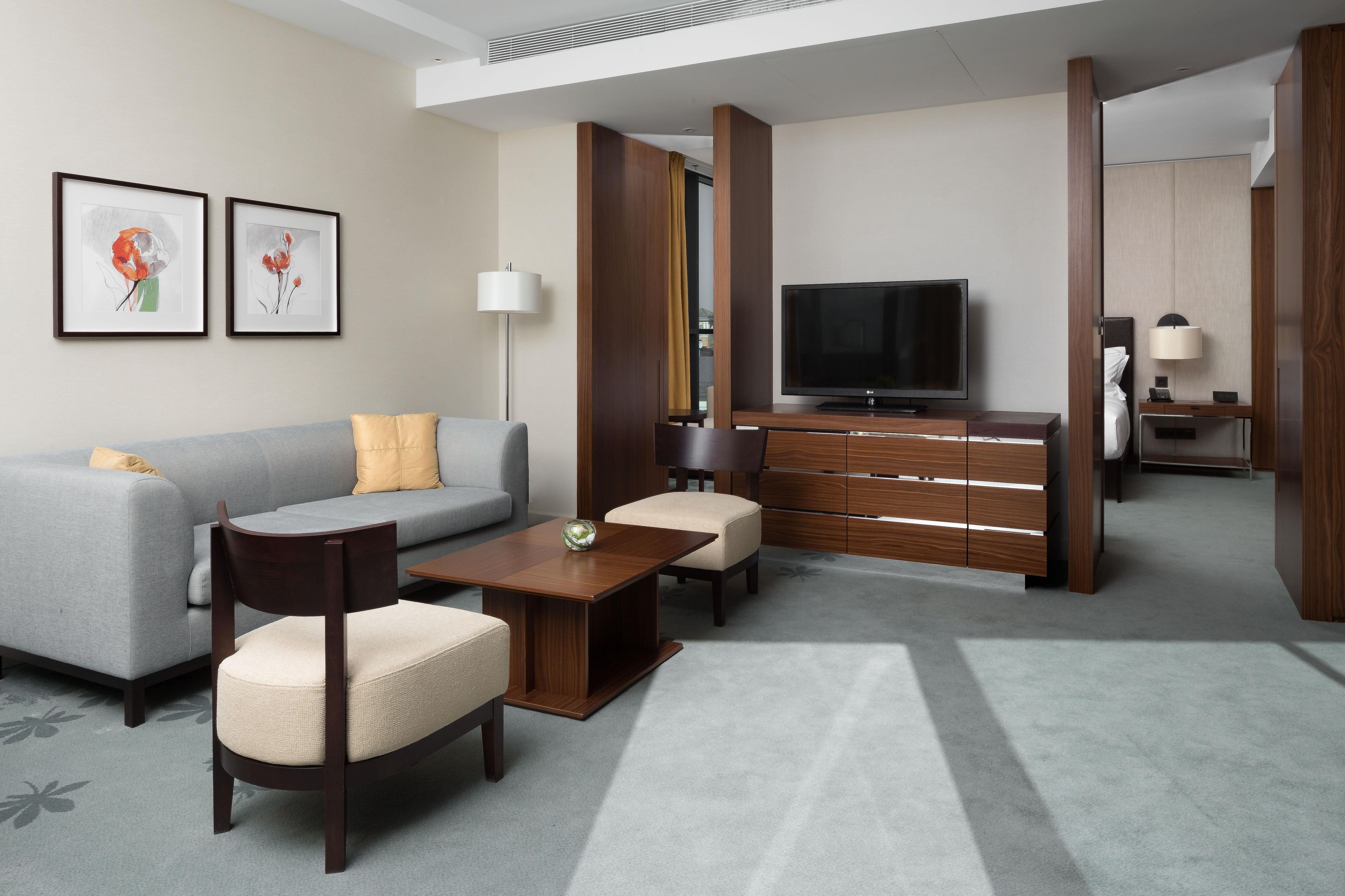 A modern hotel room with a grey sofa, a wooden coffee table, a TV, and a desk with a lamp.