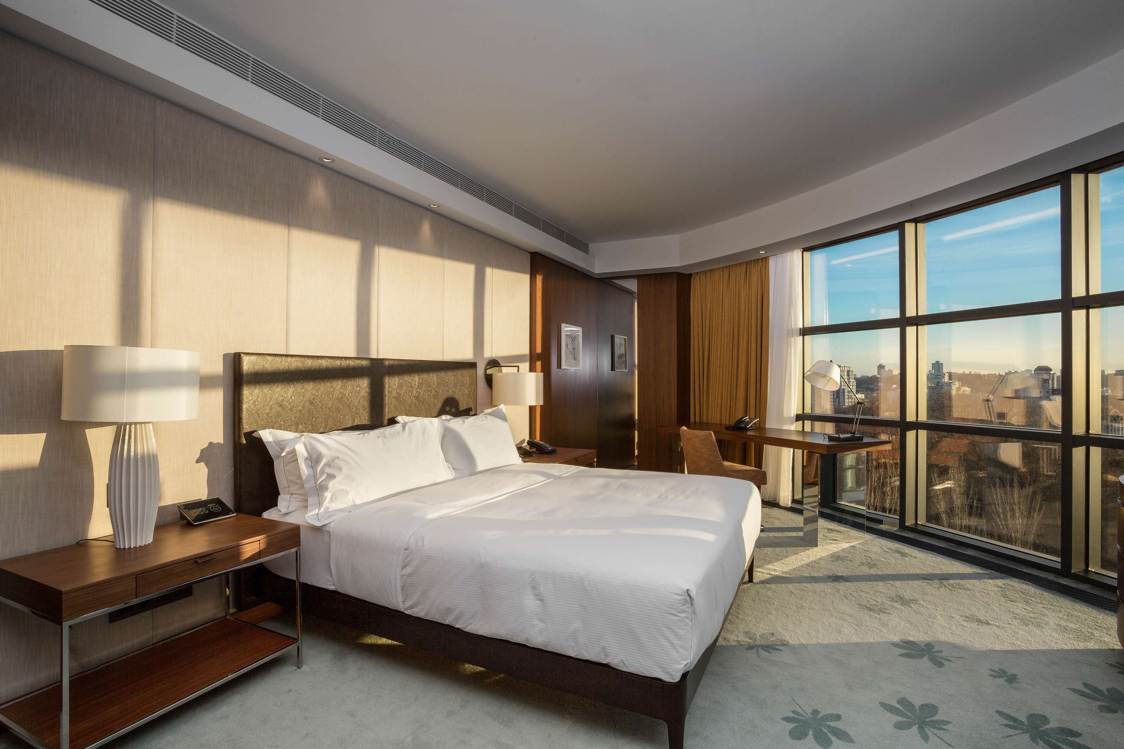 Een moderne hotelkamer met een groot bed, nachtkastjes, een bureau en stadszicht door grote ramen.