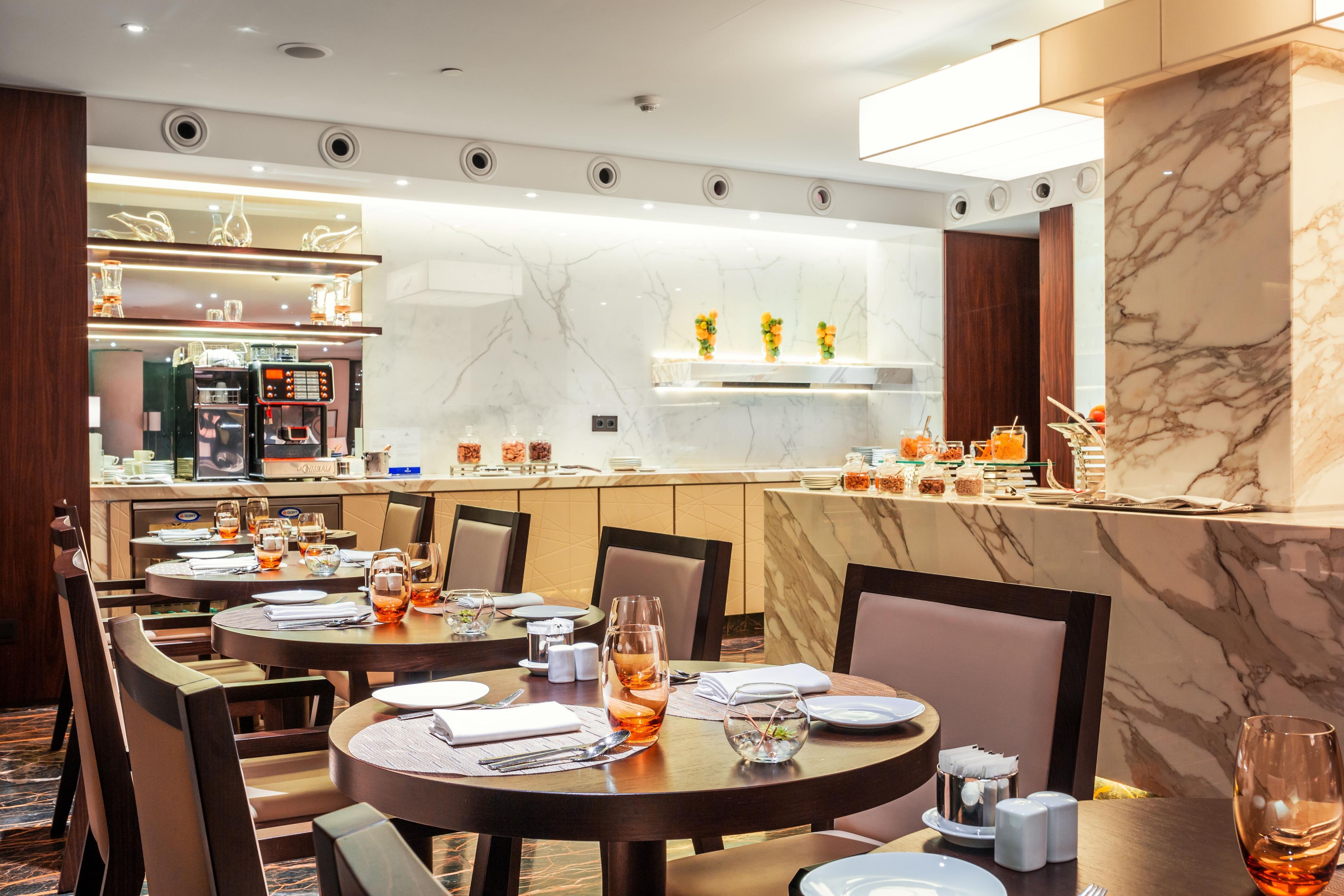 A hotel dining area with round tables, chairs, and a bar area with a coffee machine.