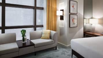 Una habitación de hotel con cama, sofá, una mesita, lámpara y ventana con cortinas.