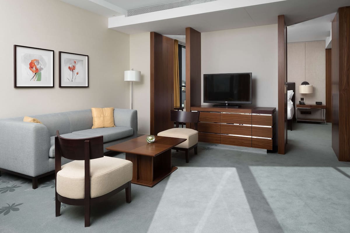 Ett modernt hotellrum med en grå soffa, ett träbord och en tv. På väggen hänger två inramade konstverk, och det finns en trävägg som avskärmning.