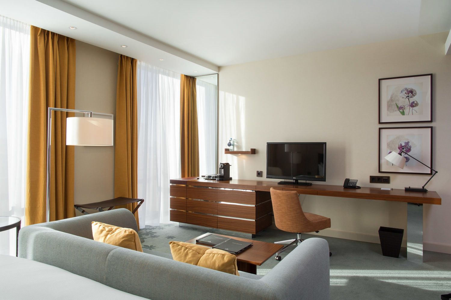 A modern hotel room with a sofa, a desk with a flat-screen TV, a lamp, and artwork on the wall.