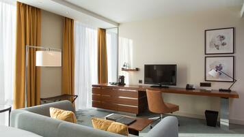 A modern hotel room with a sofa, a desk with a flat-screen TV, a lamp, and artwork on the wall.