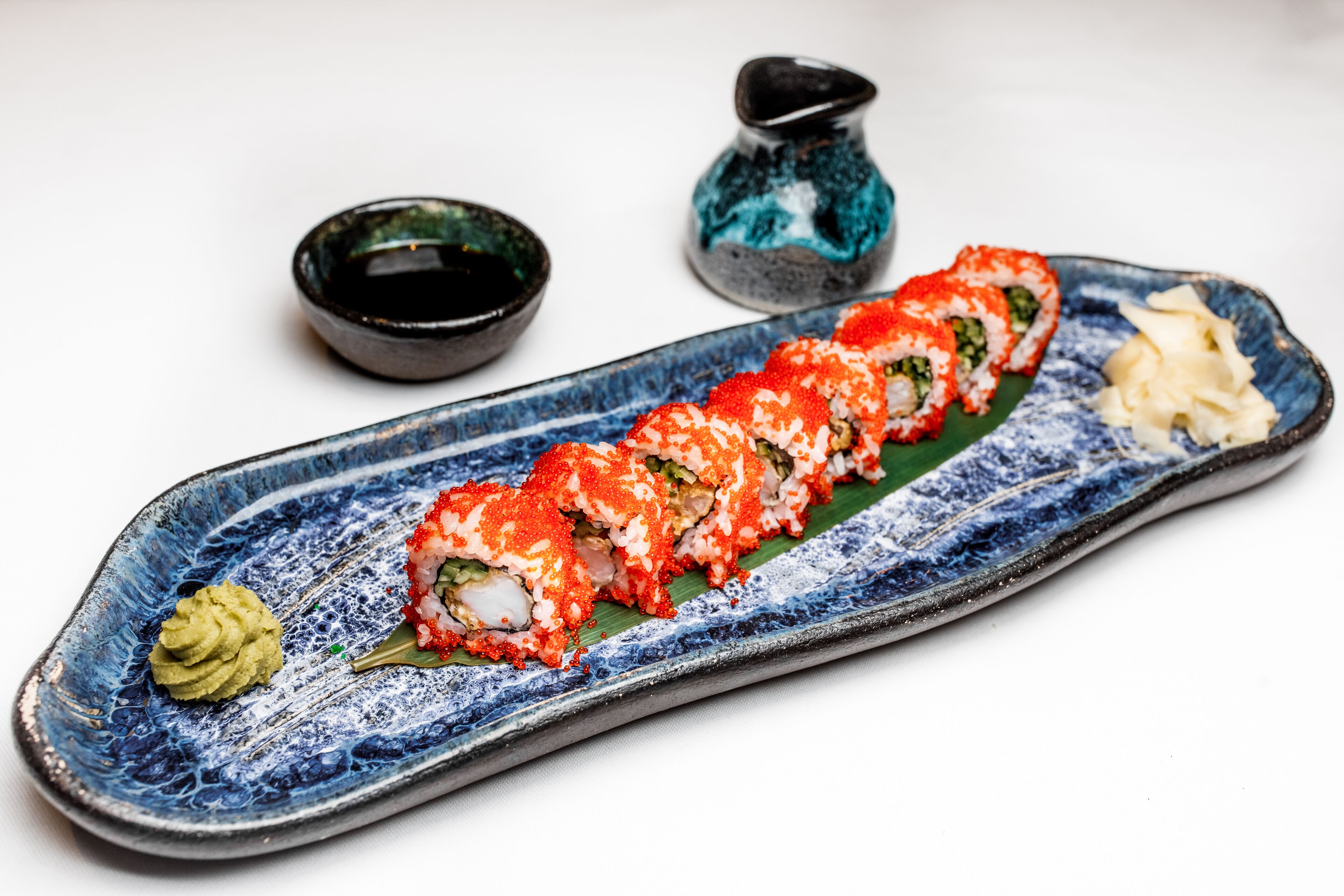 Un plato de sushi con un plato cerámico azul, un cuenco pequeño verde y dos cuencos pequeños negros con un motivo azul.