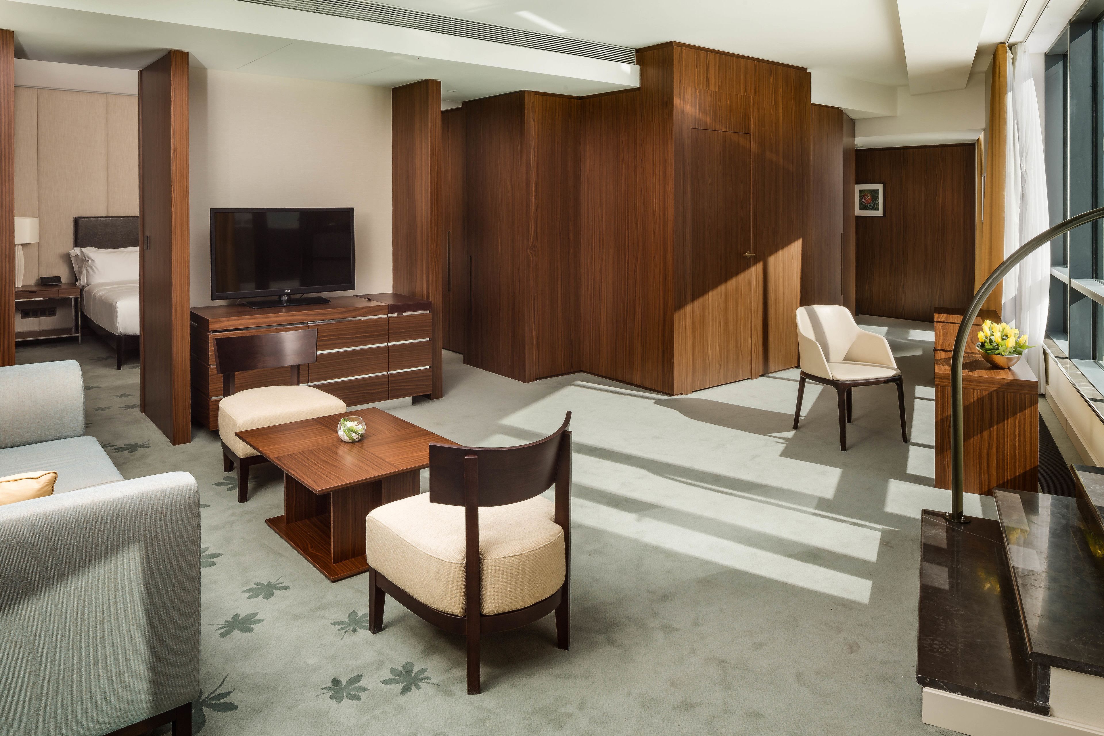 Ein modernes Hotelzimmer mit einem großen Bett, einem Schreibtisch mit Flachbildfernseher, einer Sitzecke mit Sofa und Sesseln sowie einem Holzschrank.