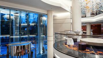 Un moderno vestíbulo de hotel con un muro de cristal que refleja el exterior, una barandilla de cristal y una lámpara decorativa.