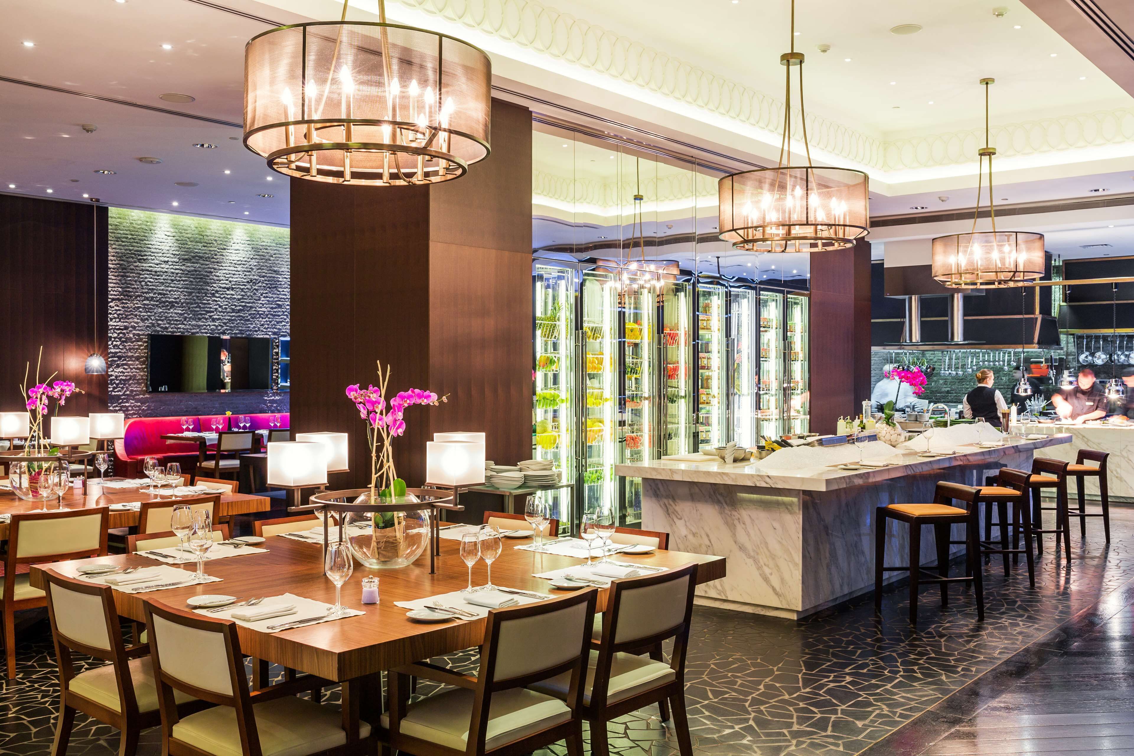 Een modern restaurant met houten tafels, hangende pendantlampen en een bar in het midden.
