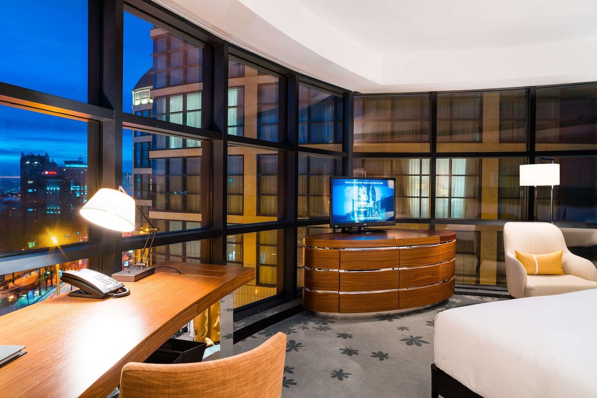 Ett hotellrum med ett stort fönster, ett skrivbord med en lampa, en tv, en säng och en stol.