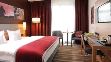 A hotel room with a large bed, a desk with a chair, a small table, and a window with curtains.