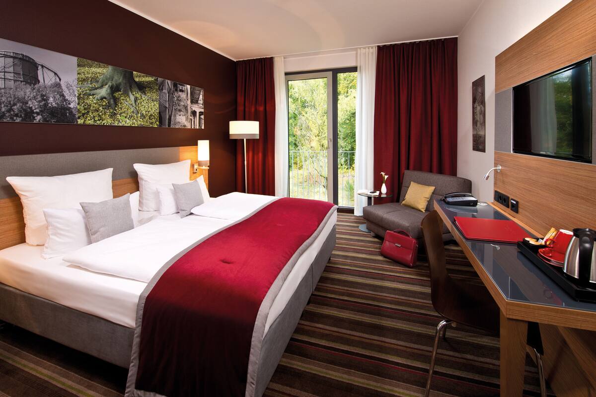 Quarto de hotel com cama grande, escrivaninha, cadeira, televisão e varanda com cortinas.