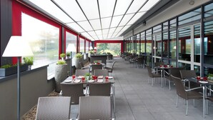 An outdoor dining area with tables and chairs, potted plants, and a view through large windows. - Leonardo Hotel Völklingen - Saarbrücken (Voelklingen)