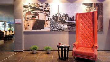 Un hall d’entrée agrémenté d’une grande fresque murale sur le thème industriel, d’un fauteuil rouge recouvert de tissu, de deux plantes en pot et d’une petite table sur laquelle se trouvent une tasse et un livre.